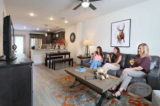 Three women sitting on a leather couch drinking wine and watching TV in a spacious apartment with hardwood vinyl floors and white walls.