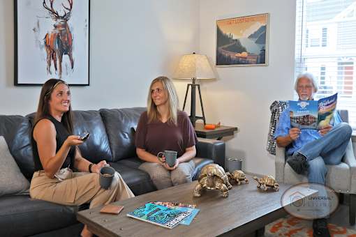 Two women sit on a couch drinking coffee and watching TV while a man sits in an arm chair reading a magazine.