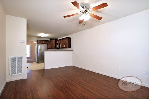 The living room with vinyl hardwood flooring, white walls, and a ceiling fan with a bar and kitchen in the background..