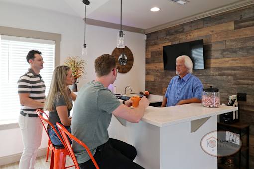 A man stands behind the coffee bar chatting with a group of people sitting at the coffee bar stool and drinking coffee.