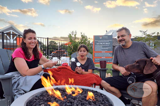 A family and their dog roast marshmallows at the firepit while the sun sets in the background.