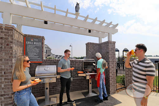 A group of people gathered around the poolside grills socializing, waiting to get burgers and drinking from orange glasses.