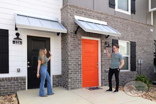 A man and woman standing outside the front doors of their apartment homes, waving at one another.