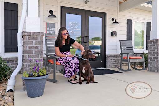 A woman sitting on a bench with her dog in front of the Clubhouse entrance.