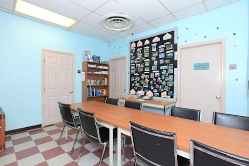 A room with a long table and chairs, a bookshelf, and a wall with pictures and a bulletin board.