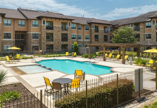 A large swimming pool surrounded by yellow chairs and umbrellas in front of a multi-story apartment building.