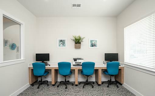 A room with a white wall and a window with blinds, has a desk with four chairs and a plant on it.