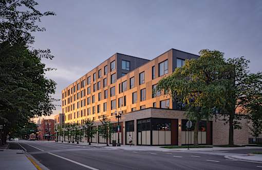 A modern building with a combination of brown and black windows is situated on a street with trees on the side.