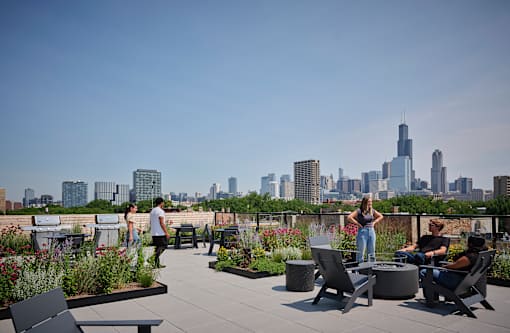 A group of people are sitting and standing on a rooftop patio with a city skyline in the background.