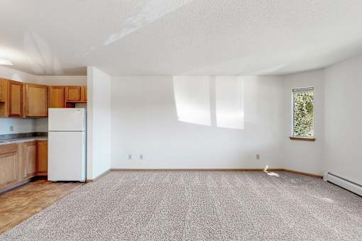 an empty living room with a kitchen with a white refrigerator