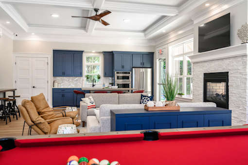 A pool table with a red cloth and white balls is in the foreground of a well-lit room with a fireplace and a television at The Promenade at Easton Apartments, Easton Maryland
