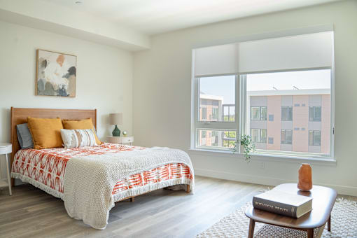 A bedroom with a bed, a small table, and a window overlooking a building.