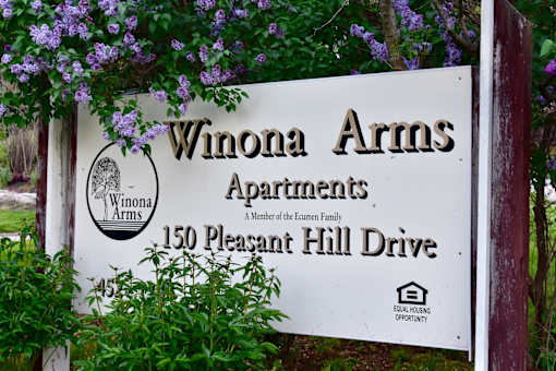 A sign for Winona Arms Apartments is displayed in front of some purple flowers.