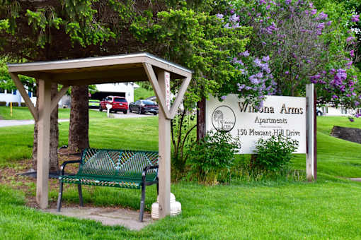 A wooden bench is situated under a wooden shelter in front of a sign for Wilona Arms Apartments.