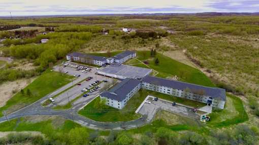 A large white building with a grey roof is surrounded by a grassy area and a parking lot.