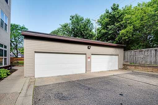 A two-car garage with a brown roof and white doors is situated in front of a building and trees.