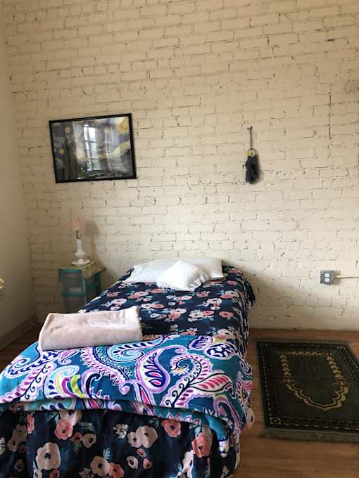 A bed with a floral patterned comforter and a pink pillow is in front of a white brick wall.