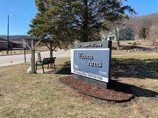 A sign for Winona Arms is displayed in front of a tree.