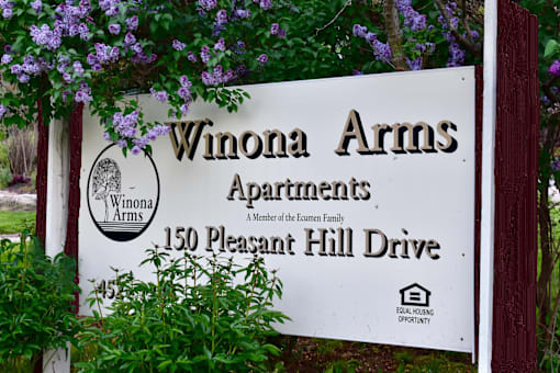 A sign for Winona Arms Apartments is displayed in front of some purple flowers.