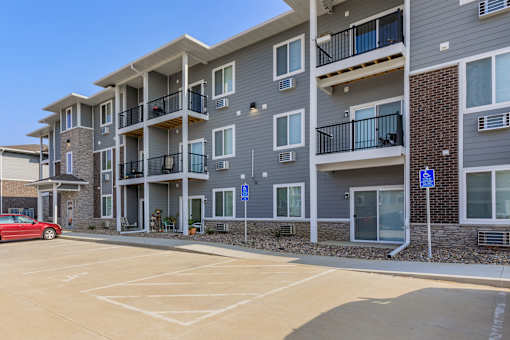 A red car is parked in a parking lot in front of a grey apartment building.