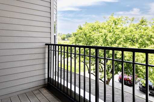 A balcony with a black railing overlooks a street with cars and greenery.