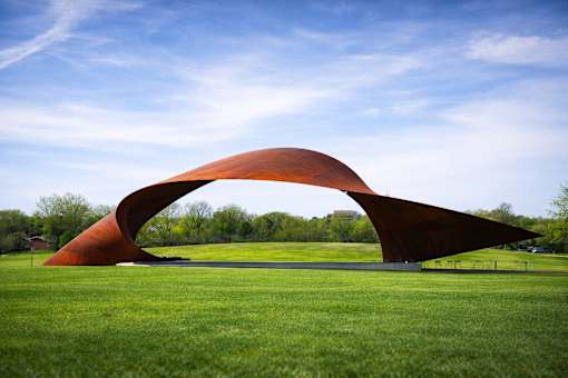 A large, curved, orange sculpture sits in a grassy field.