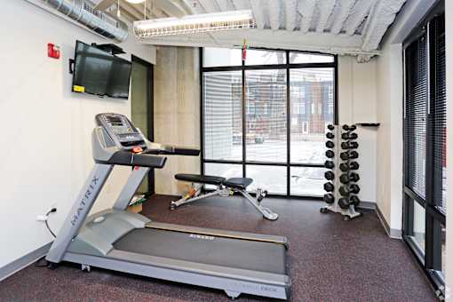 A treadmill with a TV on the wall and a window with blinds.