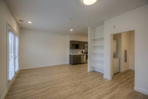 A large empty room with wooden flooring and a kitchen area in the background.