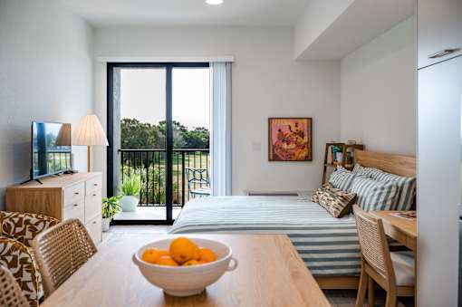 A bowl of oranges sits on a wooden table in a well-lit room with a bed and a television.at The Teale Golden Gate in Naples, FL, 34116
