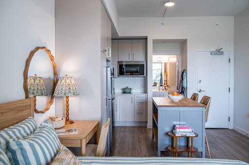 A bedroom with a bed, a desk with a chair, and a kitchen area with a refrigerator, microwave, and cabinets.at The Teale Golden Gate in Naples, FL, 34116