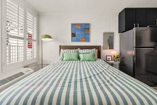 A bed with a blue and white striped comforter sits in a room with a painting on the wall and a refrigerator to the right.
