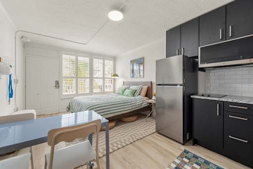 A kitchen with a refrigerator, a dining table with chairs, and a bed with a striped comforter.