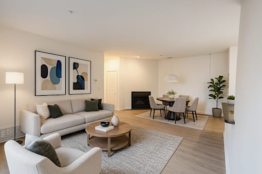 A living room with a grey sofa, a grey armchair, a wooden coffee table, a grey rug, a black fireplace, a dining table with chairs, and two framed artworks on the wall.