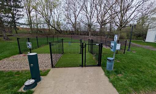 A black fence surrounds a green trash can and a grey concrete pathway.