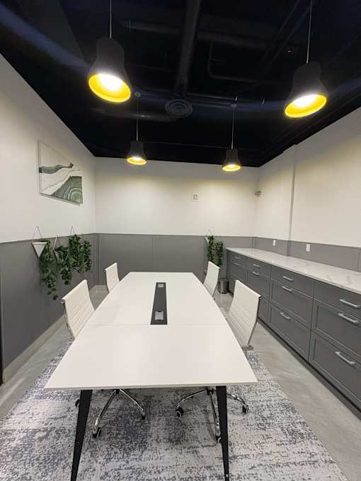 A white table with four chairs and a black object on it is in the center of a room with grey cabinets and a grey carpet.