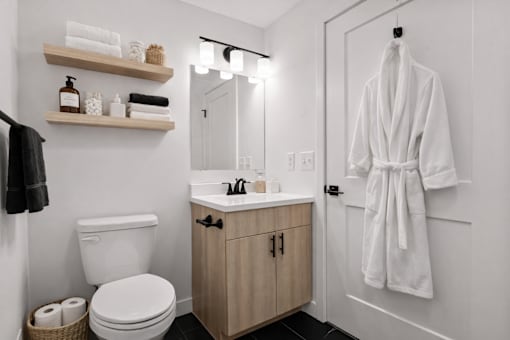 A white bathroom with a toilet, sink, and a white robe hanging on the door.