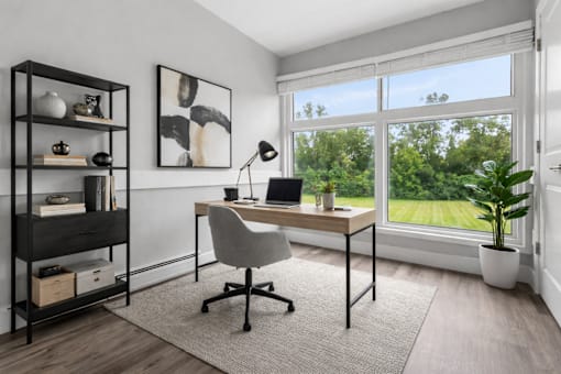 A room with a desk, chair, and a large window showing greenery outside.