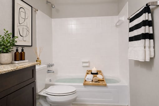 A modern bathroom with a white toilet, a bathtub with a tray on it, and a black and white striped towel hanging on the wall.