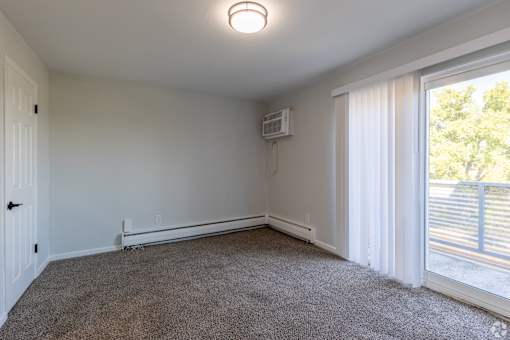 A room with a carpeted floor, a window with white curtains, and a sliding glass door leading to a balcony. at The Park on 13 Apartments - Royal Oak, MI Apartments, Royal Oak, 48073