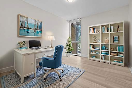 A room with a blue chair, a desk with a laptop, a bookshelf with books, and a painting on the wall.