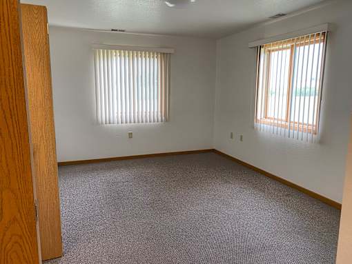 A room with a carpeted floor and two windows with blinds.