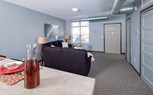 A room with a bed, a couch, a table with a bottle and a plate of food on it, and a window with a view of the outside.
