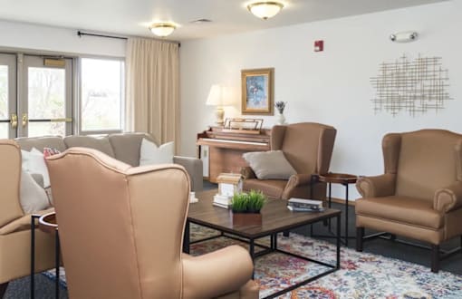 A living room with a brown chair in the foreground and a white wall with a picture frame and a fire alarm.