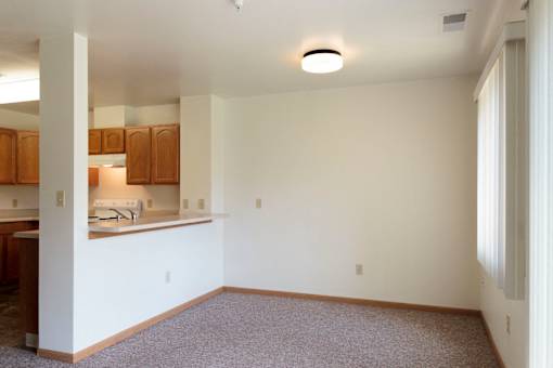A room with a carpeted floor and a kitchen area with wooden cabinets.