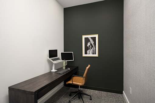 A black wall with a framed picture and a desk with a laptop and a monitor.