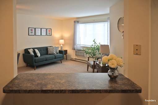 A living room with a dark green couch, a coffee table with a vase of flowers, and a window with white curtains.