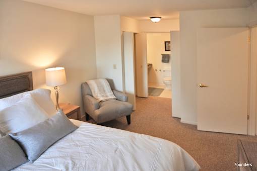 A bedroom with a bed, chair, and bathroom visible through the door.