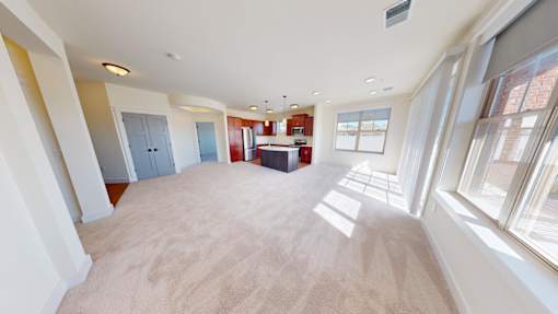 A large, empty room with a carpeted floor and a kitchen area in the background.