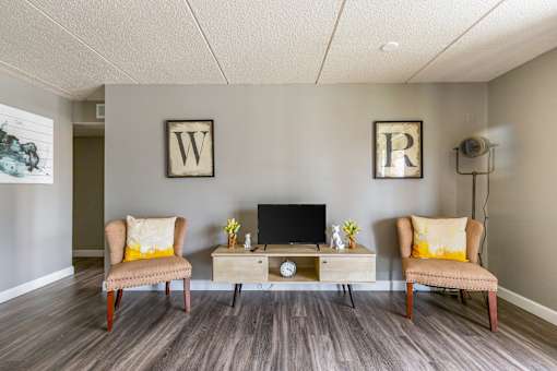 A room with a wooden floor, two chairs, a table with a laptop and a clock.
