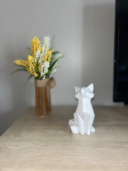 A white ceramic fox figurine sits on a wooden surface next to a vase with yellow flowers